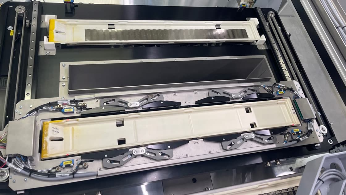 Photo of the real machine from above: two workpiece carriers, one with a row of foil strips, an empty holder between them, surrounded by clamping levers, sensors and a linear axis.