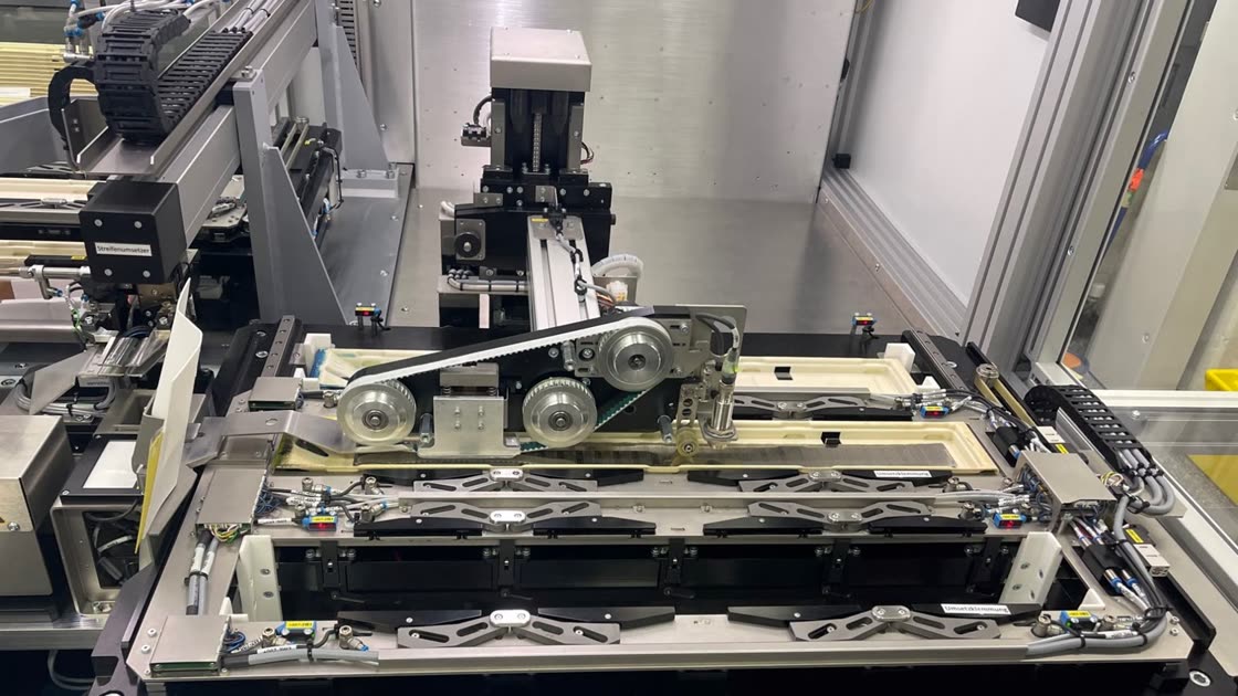 Photo of the real machine: a belt head with pulleys above a workpiece carrier with foil strips, in front the levers of the transfer clamp, behind it the gantry of the strip transfer unit.