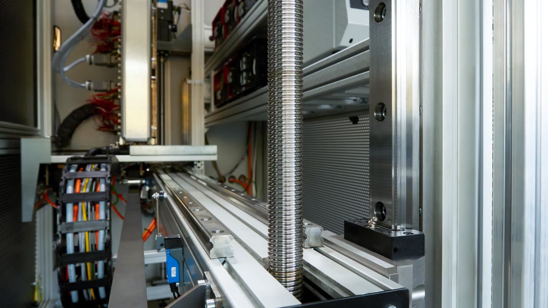 Photo from inside the system: a lead screw with bellows, linear guides and a cable carrier full of cables, on the left the sensor fixture with red sensor cables, in the background the radiant heaters in rows.