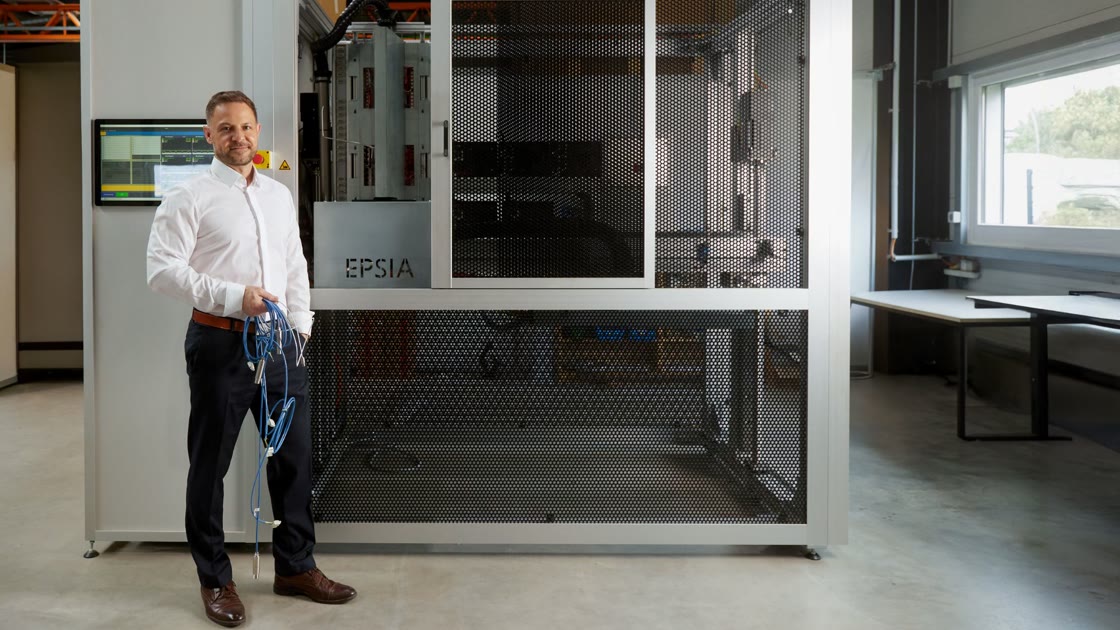 Photo of the finished calibration system in a hall: a silver-grey enclosure with screen, emergency stop and perforated sheet-metal doors, behind the open door the sensor fixture with the EPSIA lettering; in front on the left a man in a white shirt holding sensors on blue cables.