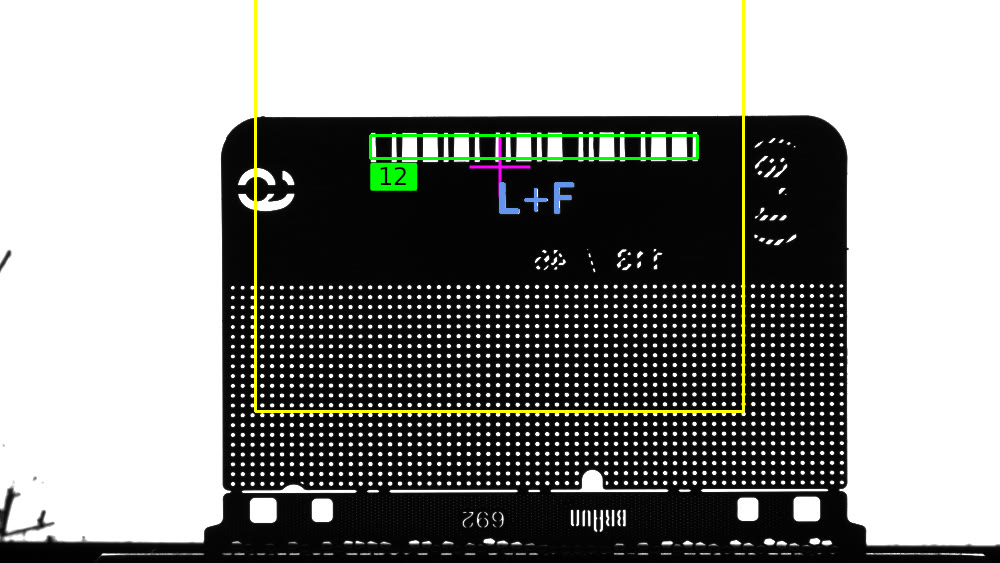 Black-and-white camera image of a foil strip in front of a light panel: a yellow search area, a line marking framed in green with the number 12 and the identifier L+F overlaid in blue.