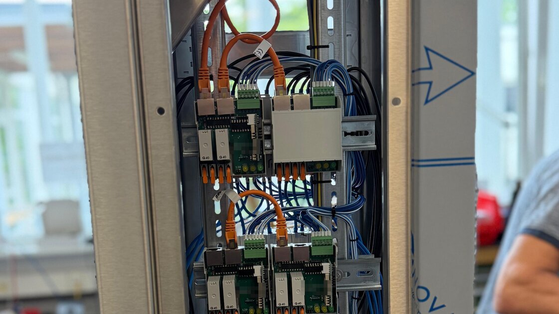 View into an open pedestal: several green bus modules with relays and LEDs on DIN rails, connected by orange patch cables for the RS-485 bus, behind them blue control wires; the manufacturer's print on the boards is obscured.