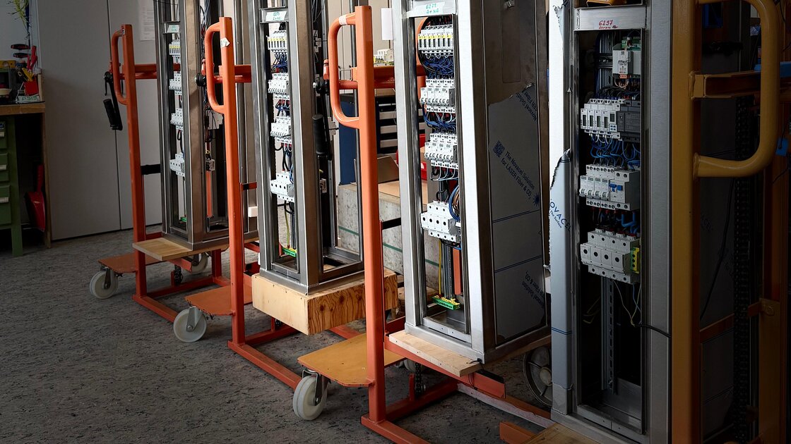 Five stainless steel supply pedestals being assembled in a workshop, each open on a hand truck: rows of circuit breakers, residual current devices and terminals wired in blue, with protective film on the panels.