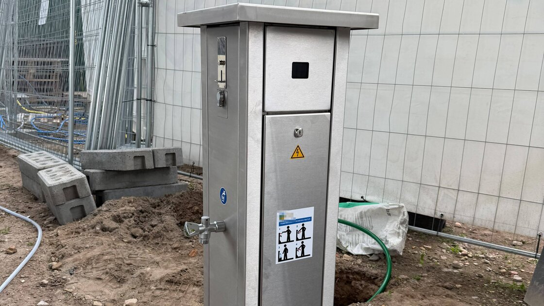 A stainless steel water pedestal being installed at a pitch, dug-up soil and hoses in front, construction fencing behind: a small keypad with display at the top, the tap on the side, a sticker on the door with drinking water instructions; the manufacturer's name is obscured.
